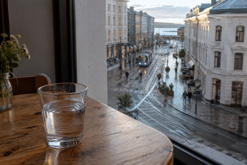 A glass tumbler of water standing on a wooden table beside a large window