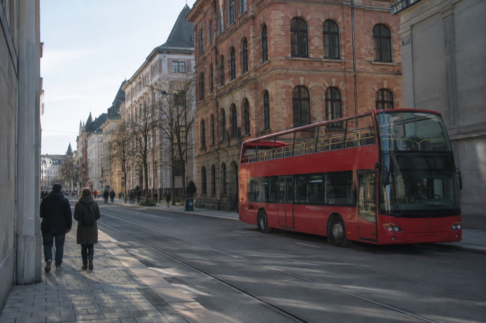 A red open-top sightseeing bus parked on a street in central Oslo with old brick buildings behind it