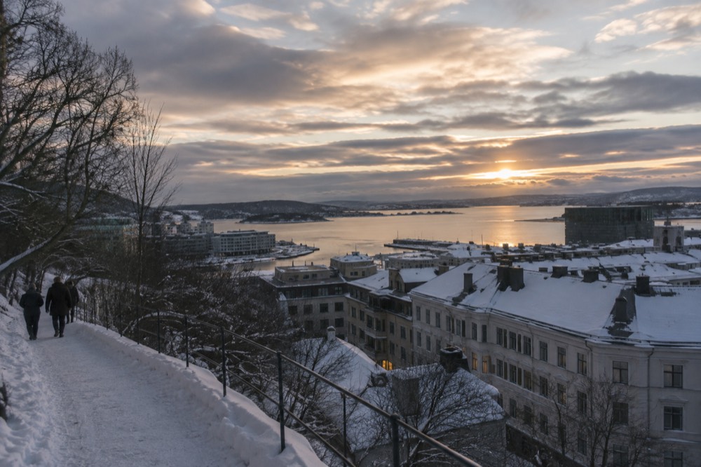 Snow-covered city rooftops seen from a hillside above a fjord at dusk