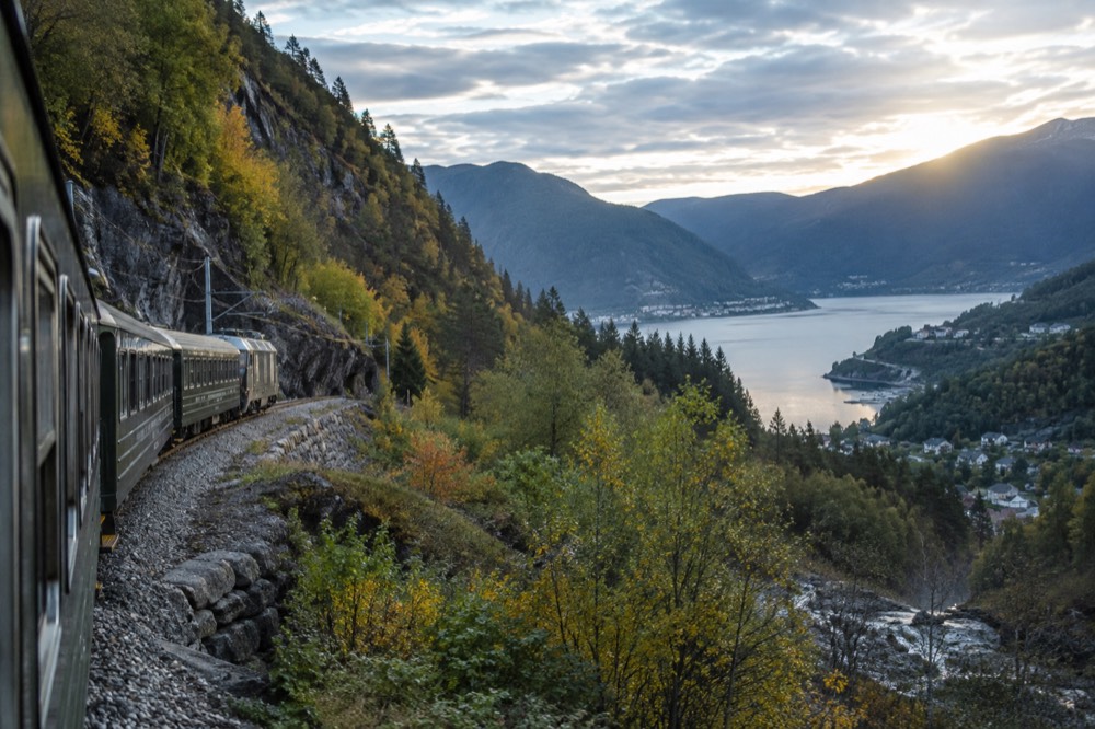 A train winding down a steep forested valley toward a fjord in western Norway