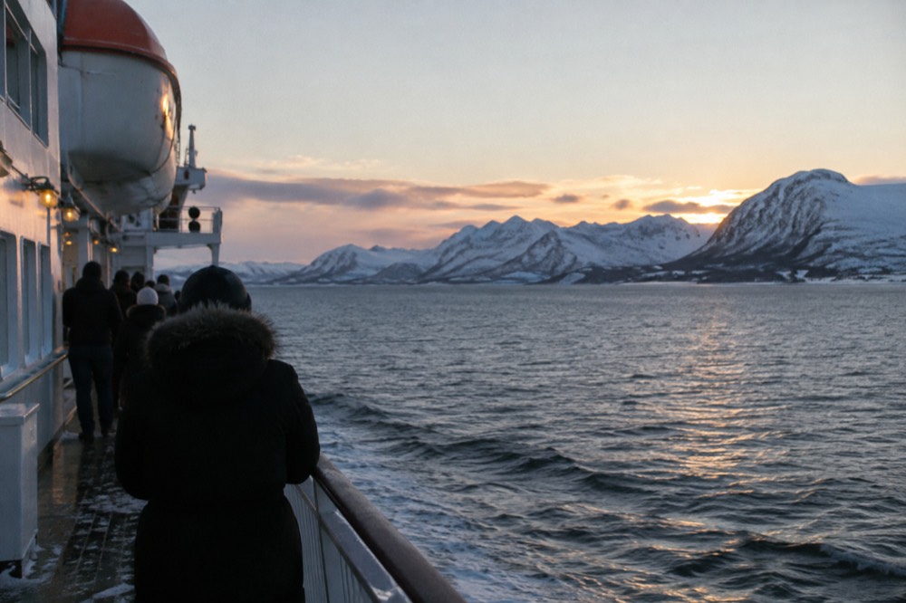 Coastal steamer ship sailing along snowy Norwegian coastline at dusk