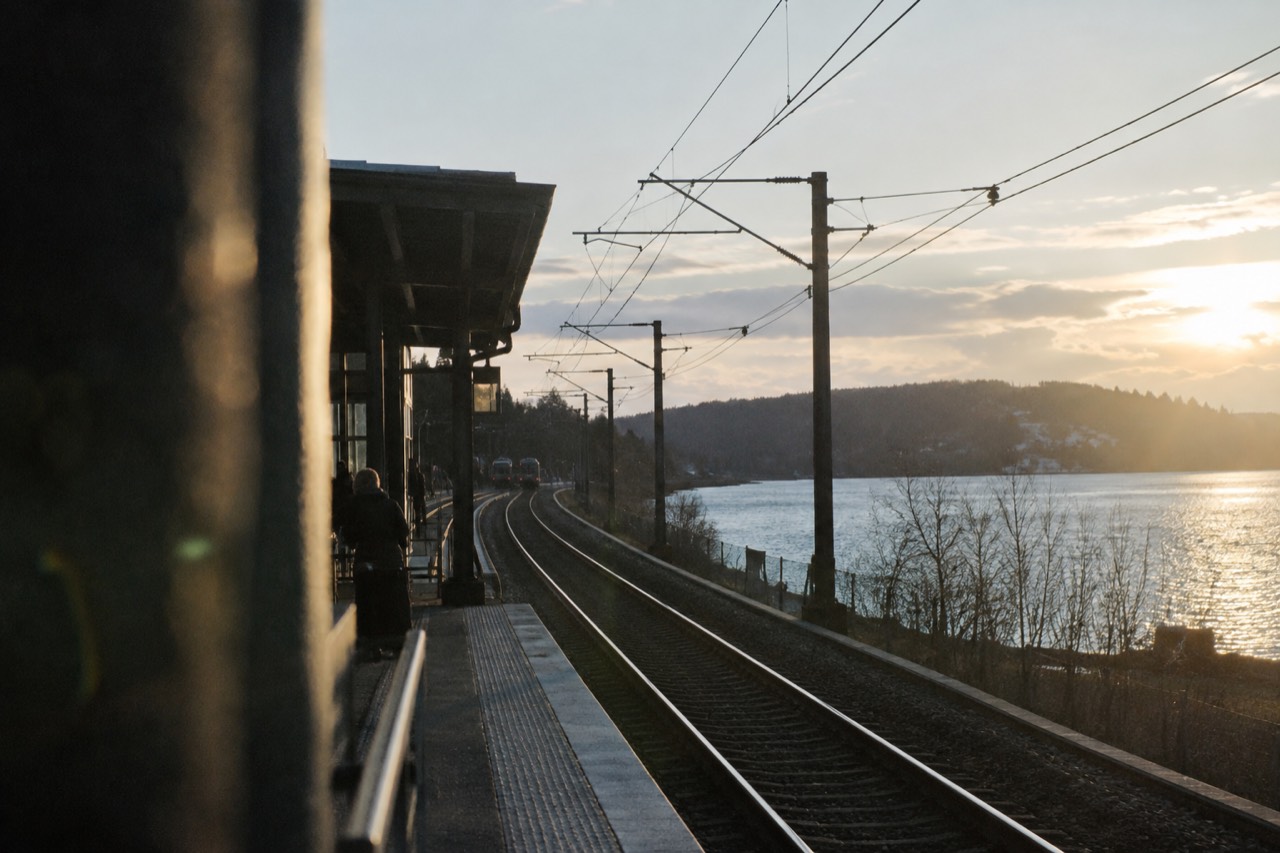 Train tracks leaving a Scandinavian station beside a fjord inlet with forested hills