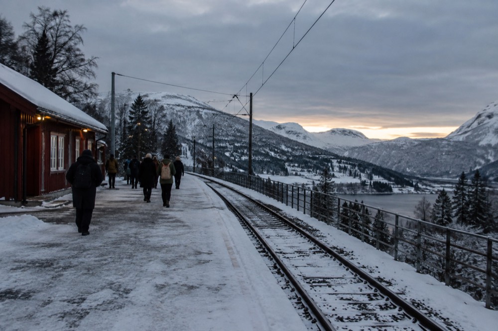 A train platform at a small mountain station under low evening light with snow on the peaks