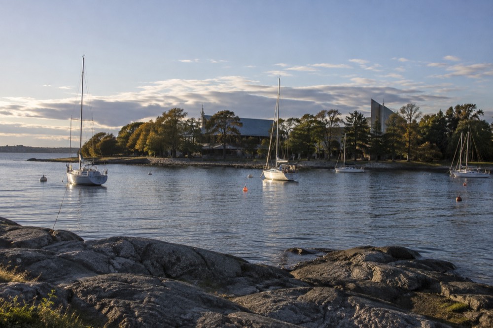 Sailboats moored off a green peninsula shoreline with museum buildings behind trees
