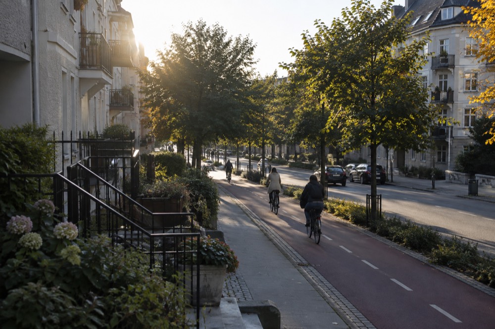 Cyclists riding a bike lane past apartment buildings and green trees on a city street
