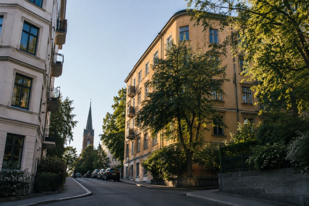 An apartment-block street corner with trees and a distant church tower