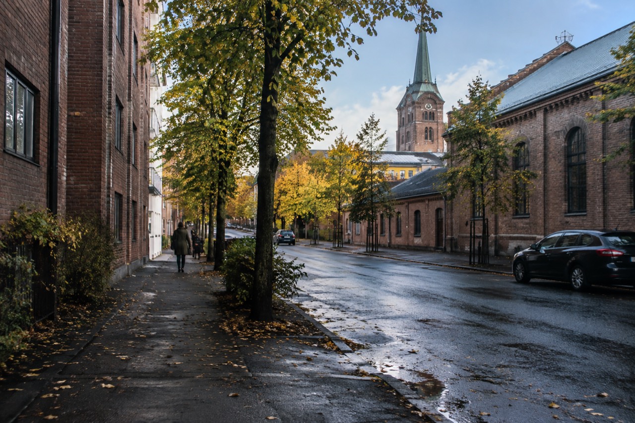 Tree-lined street with brick warehouse buildings and a church tower behind them in central Oslo