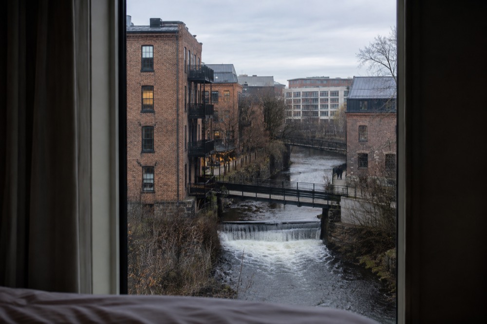 A hotel room window overlooking a river and converted brick buildings