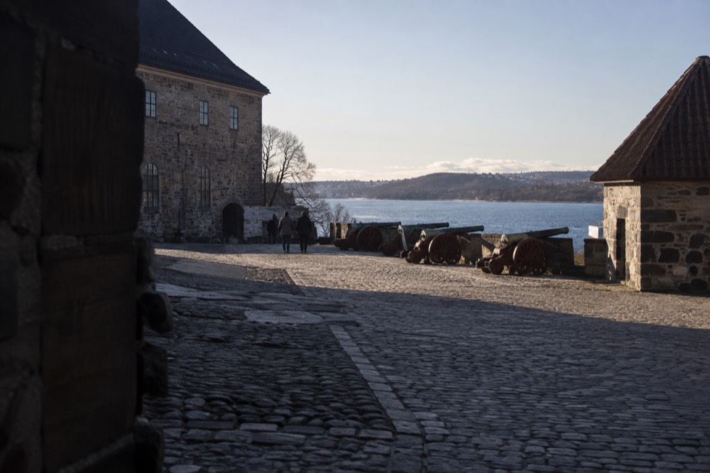 Cobblestone courtyard inside the fortress with old stone buildings and cannons facing the fjord