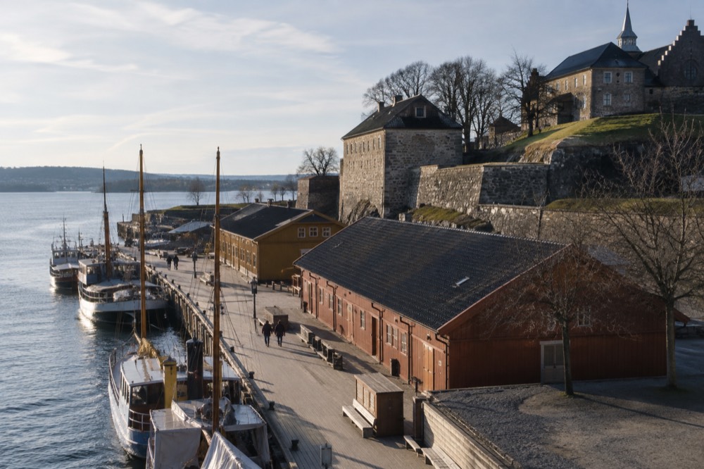 Waterfront promenade with wooden buildings, moored boats and fortress walls in the background