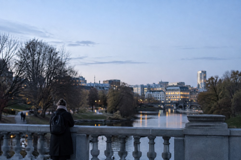A stone park bridge over a river in Oslo with the city skyline beyond the trees