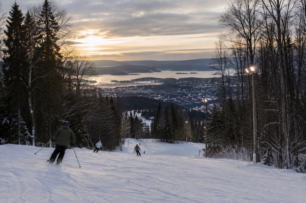 Skiers making turns down a floodlit slope with the Oslo city lights visible below the forest