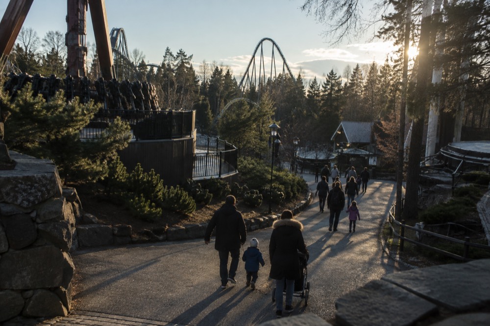 Families walking along a landscaped path between ride entrances in an amusement park