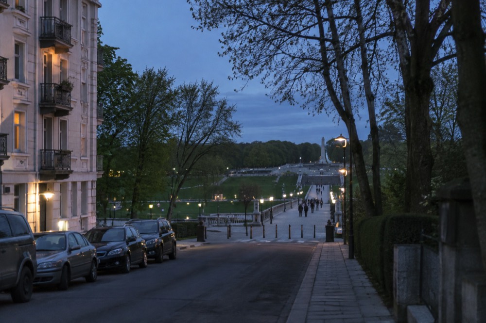 A city street with apartment buildings and trees leading toward a park entrance
