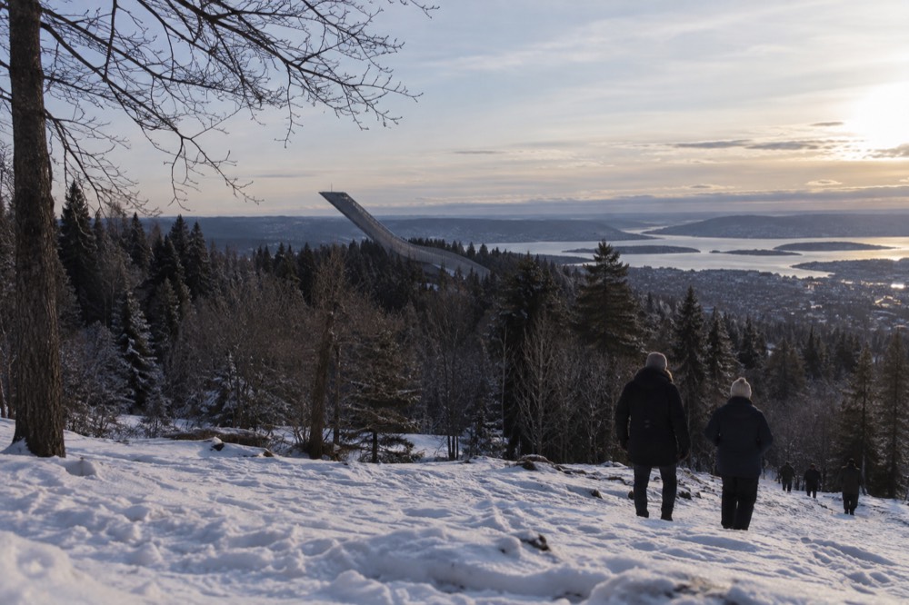 A ski jump tower above pine forest with the city and fjord visible in the distance