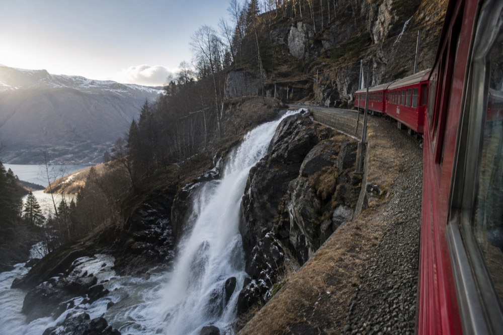 A red train on a mountain railway curving beside a waterfall and steep rock wall