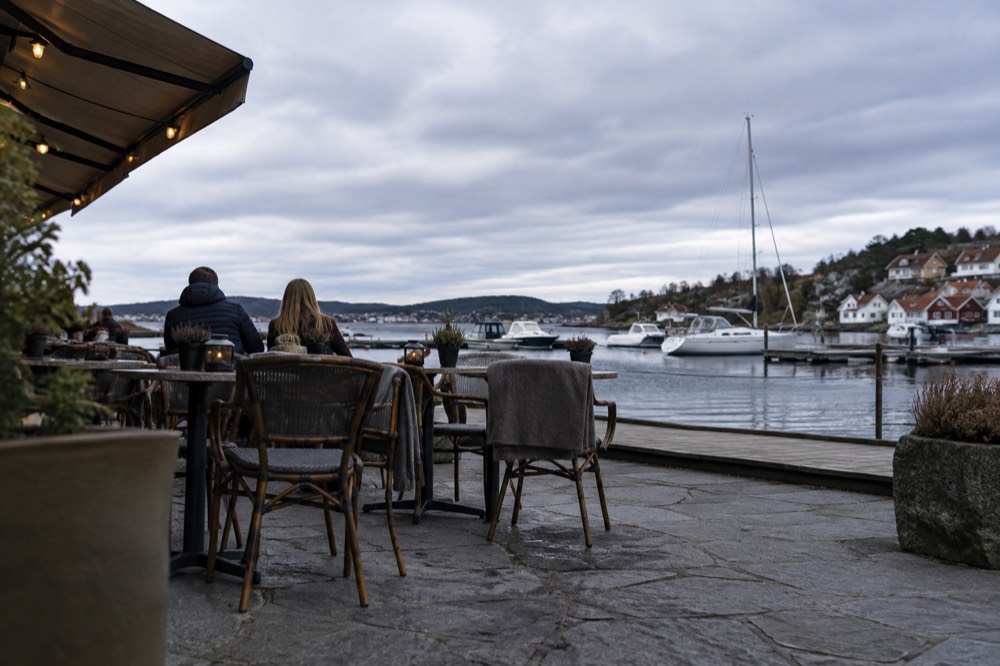 A café terrace beside a harbor with chairs facing the water and boats moored nearby