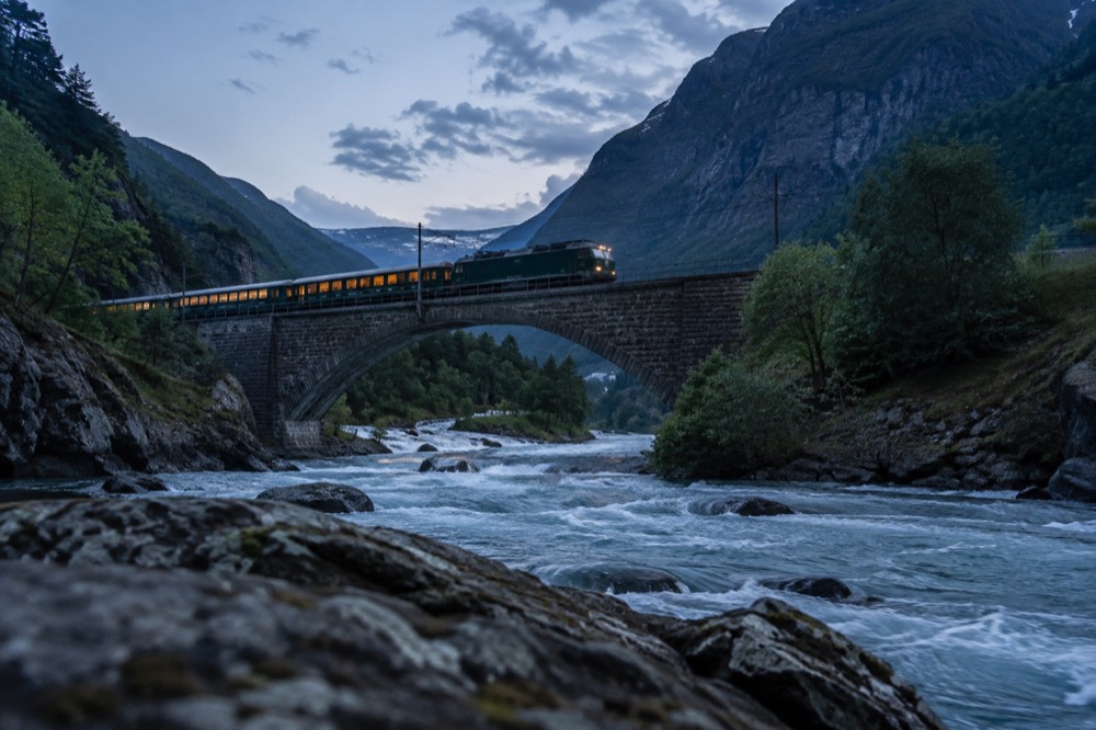 A train crossing a stone bridge over a river in a narrow mountain valley at dusk