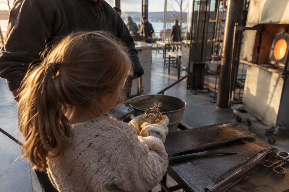 A child wearing safety gloves holding a small freshly blown glass object with an instructor nearby