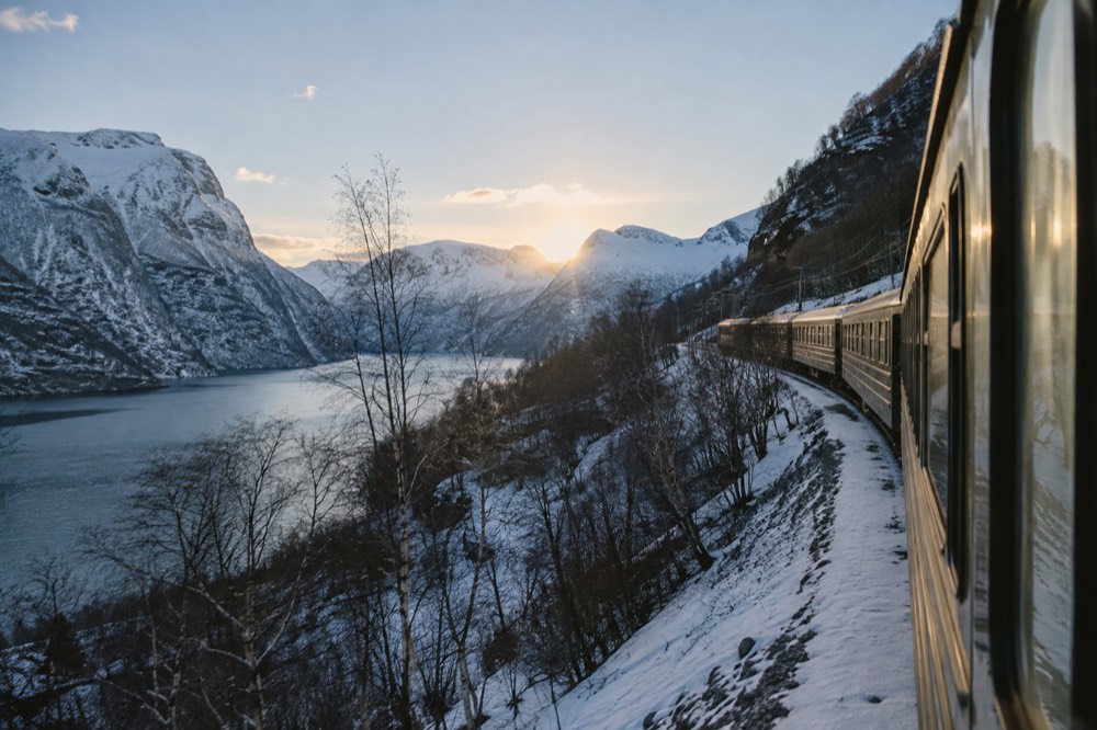 A passenger train winding along a steep fjord valley with snow-capped mountains above the water