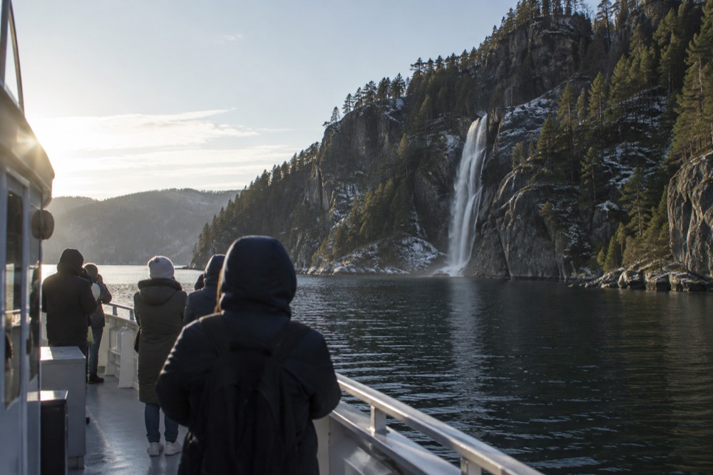 A fjord boat deck with passengers in jackets looking up at a waterfall on a cliff face