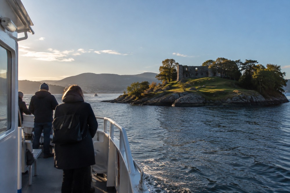 A small passenger ferry approaching a green island with a stone ruin in Oslo harbor