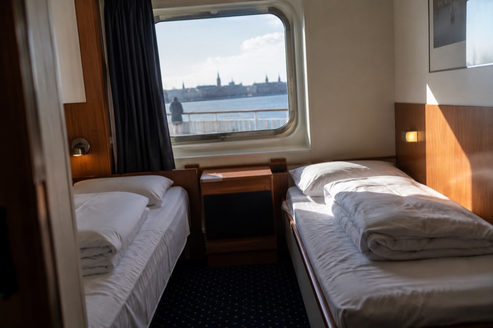 A compact ship cabin with two beds, a window, and folded white bedding