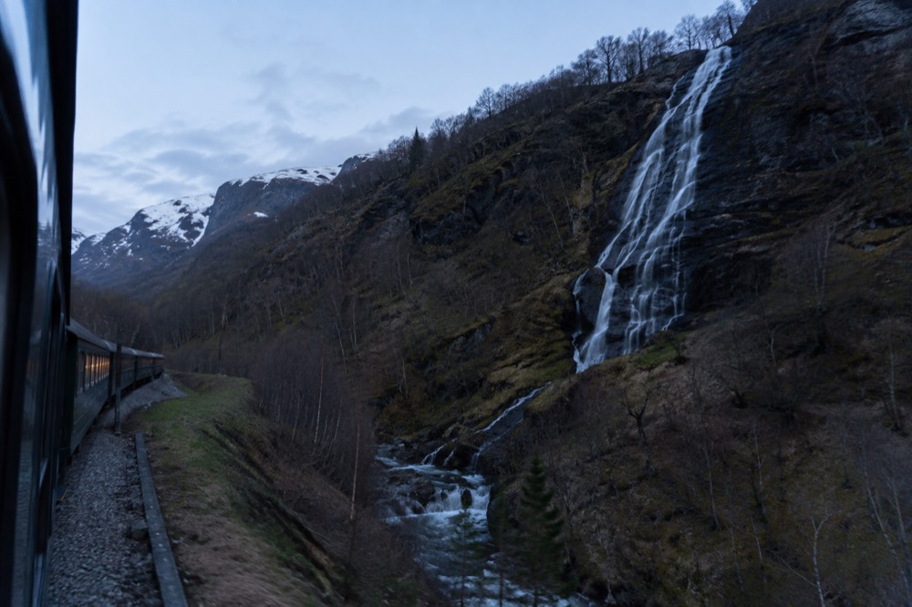 A waterfall dropping in thin white ribbons down a dark rock face beside railway tracks