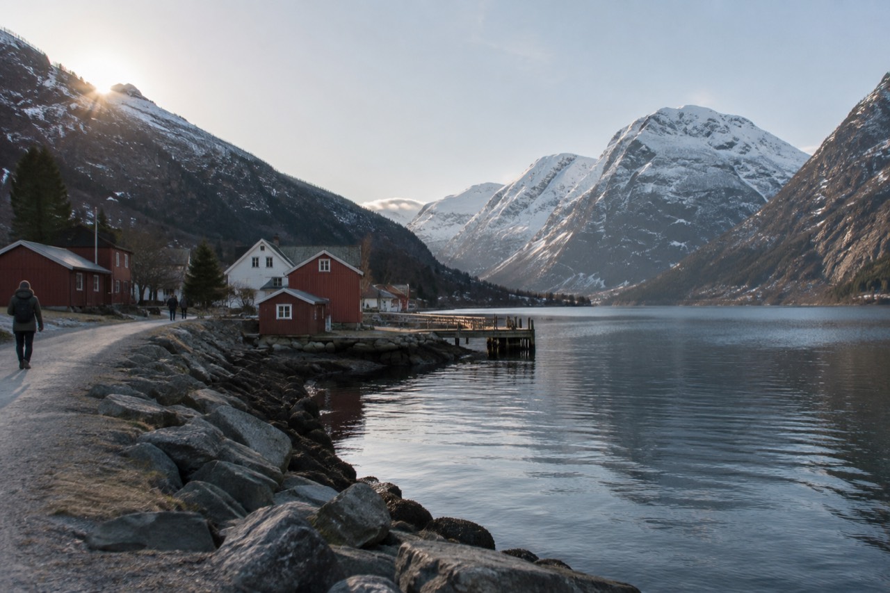 A small village of red and white buildings beside a narrow fjord with steep forested mountains rising on both sides