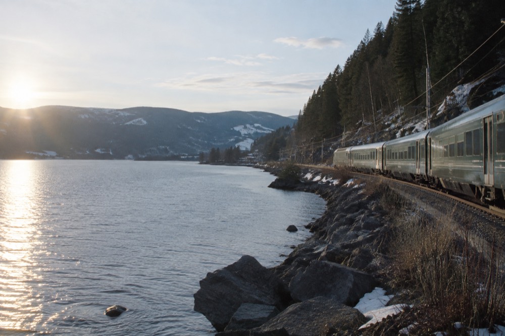 A train travelling along a shoreline with forested hills under a pale sky