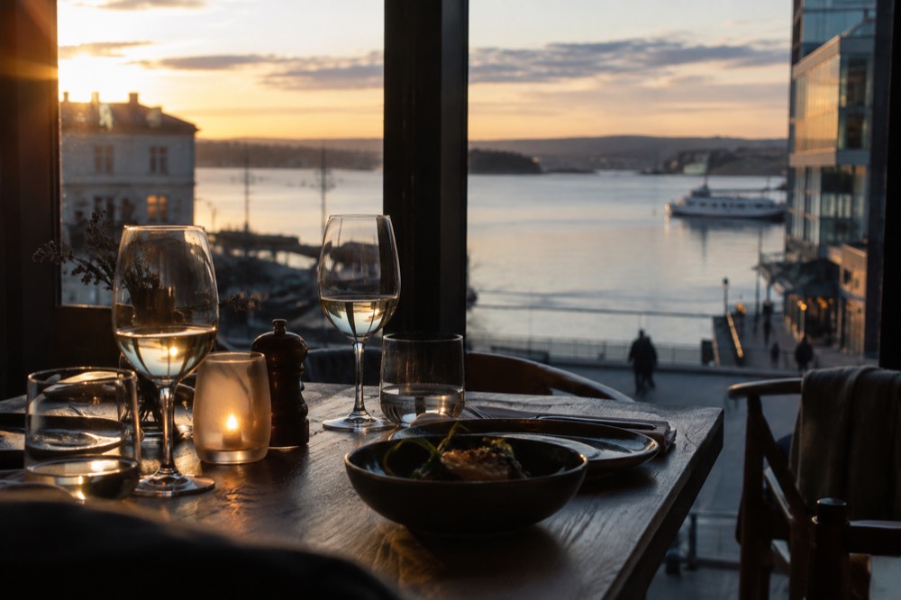 A candlelit restaurant table with wine glasses beside a window overlooking Oslo's waterfront at dusk