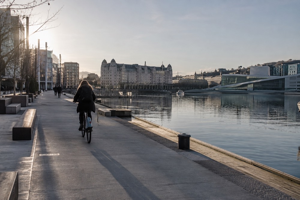 A cyclist riding a shared bike along a wide waterfront path beside calm harbor water