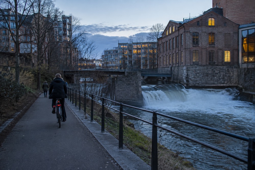 A riverside cycling path beside a small waterfall with old industrial buildings on the far bank