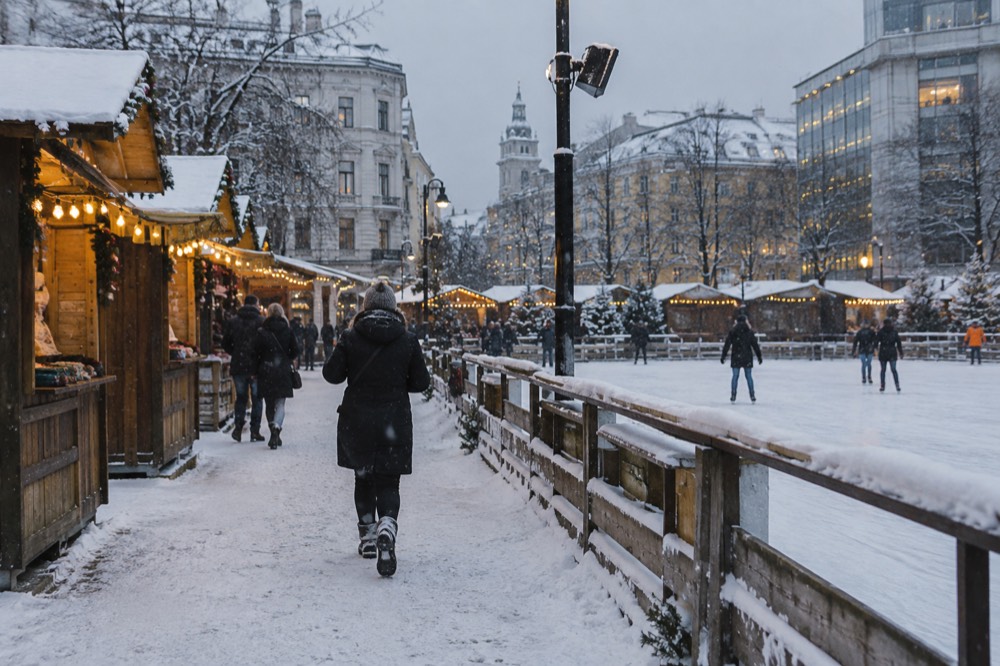 Wooden market stalls with string lights beside an outdoor skating rink in falling snow