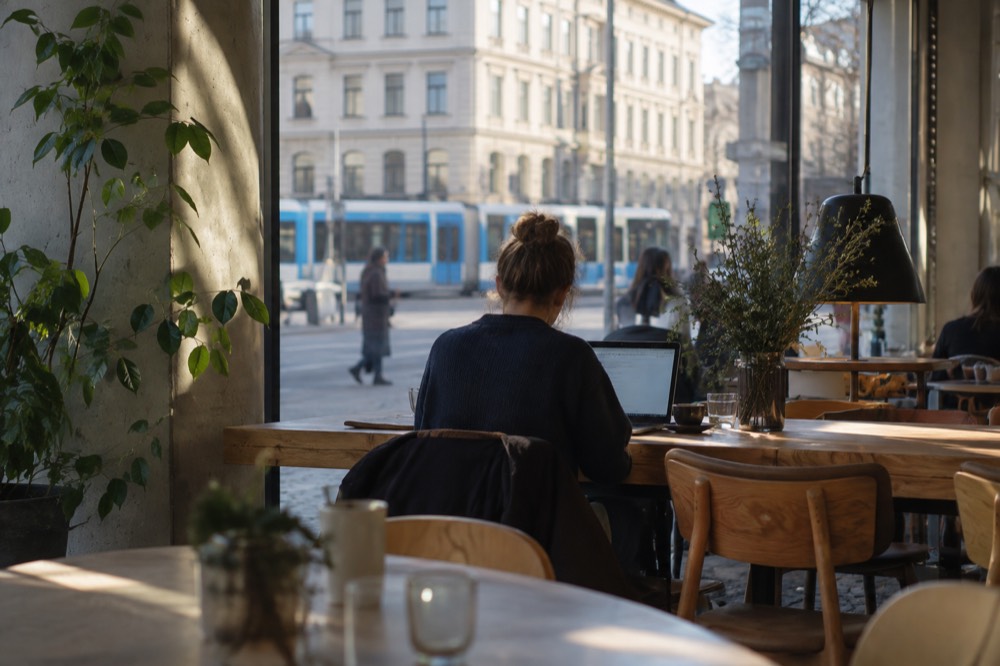 A person working on a laptop at a long shared wooden table in a bright Oslo cafe