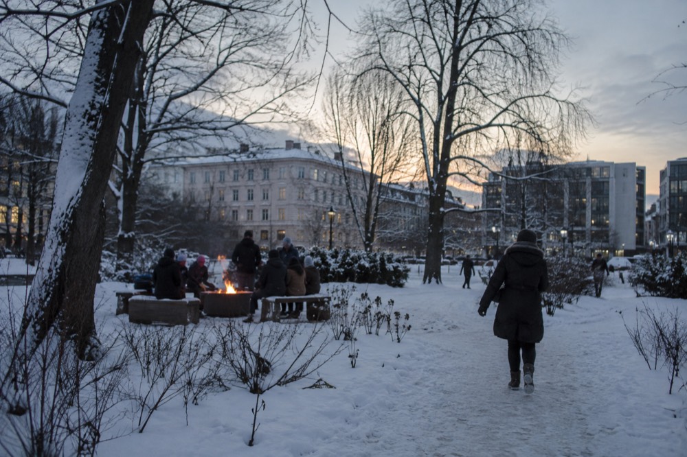 People gathered around an outdoor fire pit in a snowy Oslo park at dusk