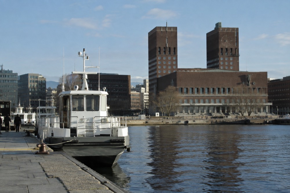 A small ferry docked at a city pier with the Oslo City Hall building behind it