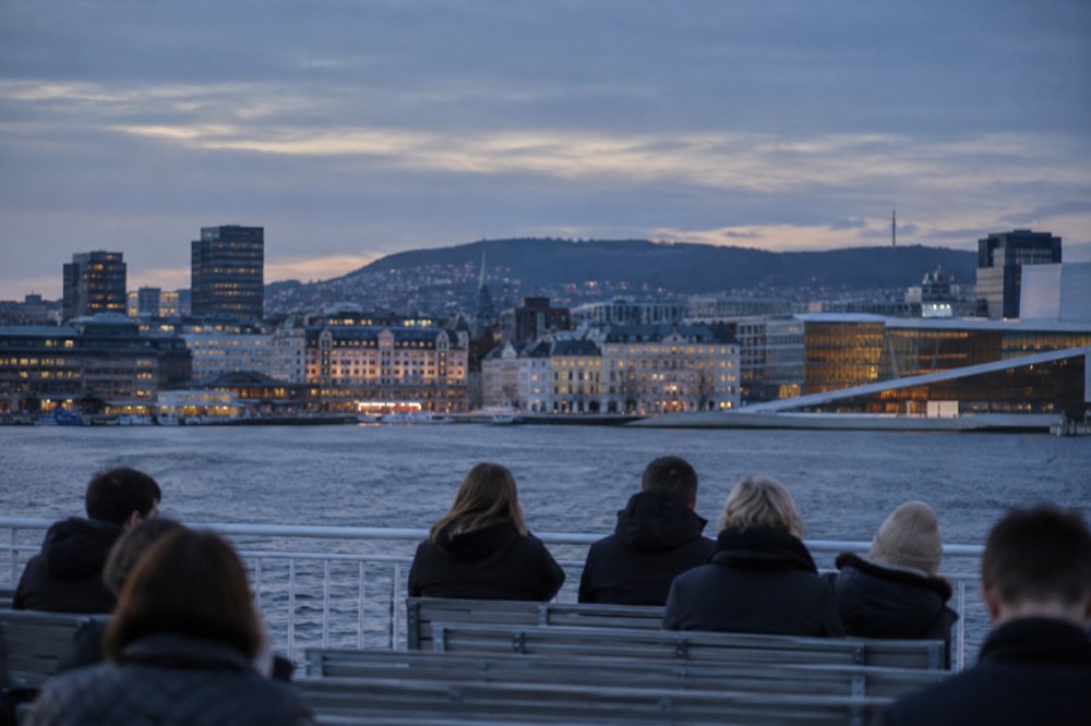 Passengers sitting on the open upper deck of a ferry with the Oslo skyline behind