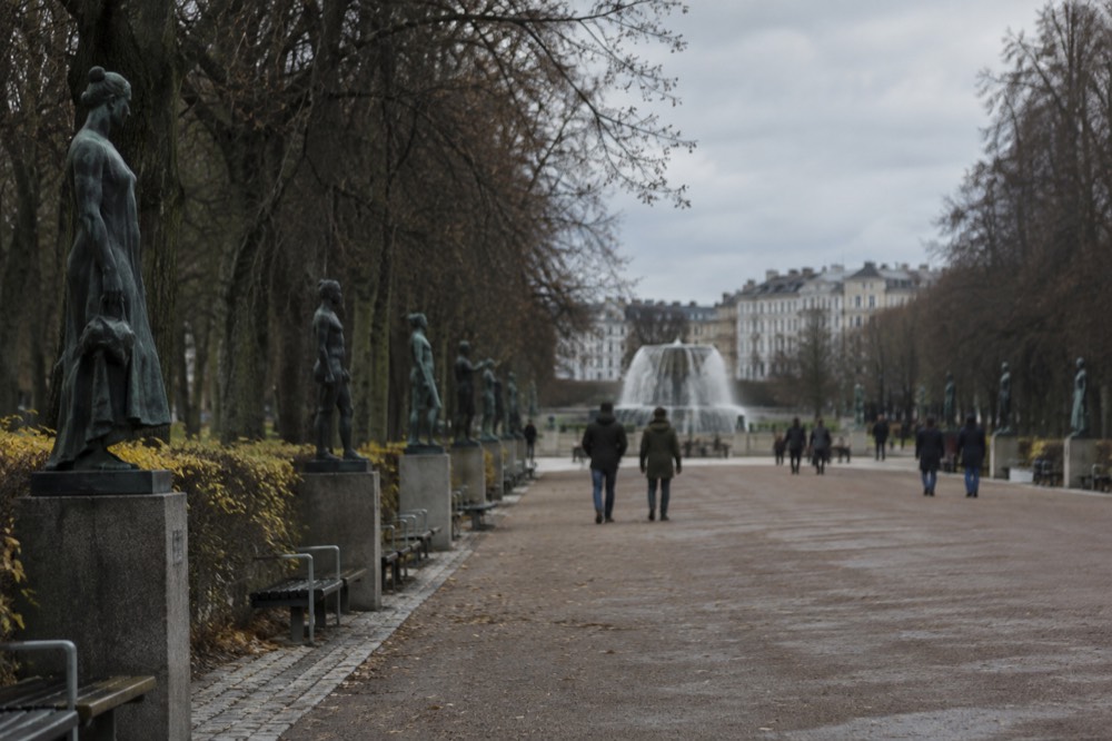 A wide sculpture park avenue lined with trees and bronze figures on pedestals leading to a fountain