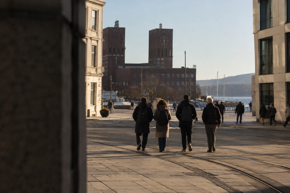 A group of pedestrians with backpacks crossing a plaza toward a large brick building with a clock tower
