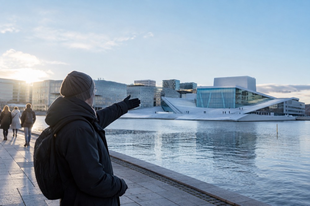 A guide gesturing toward a modern glass opera house with a sloping white marble roof beside the water
