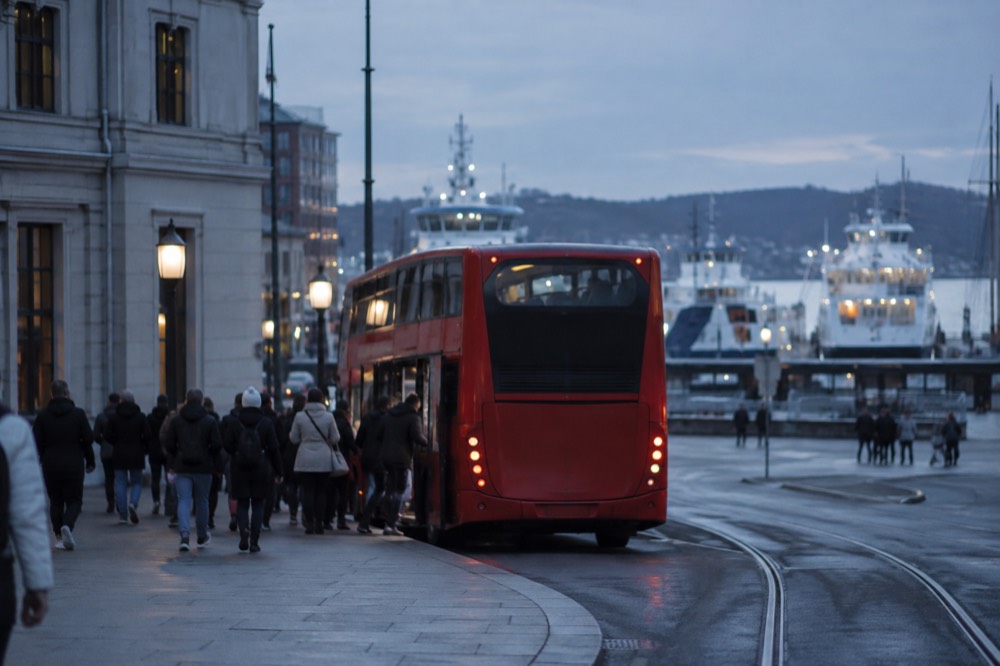 Passengers boarding a red sightseeing bus near the Oslo harbor with ferries moored behind