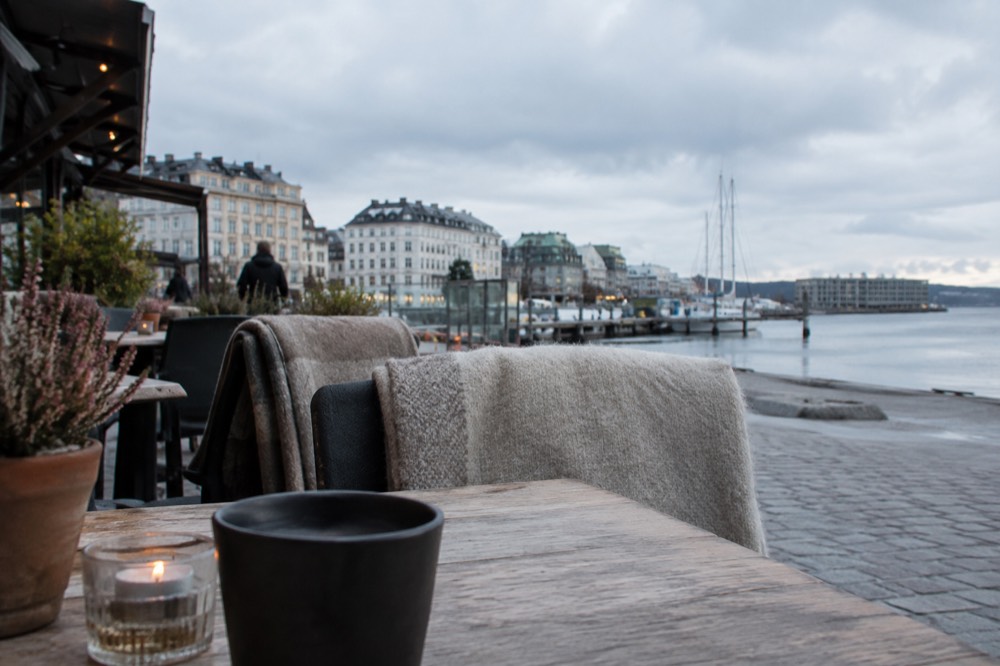 Outdoor restaurant terrace with blankets on chairs beside a fjord promenade