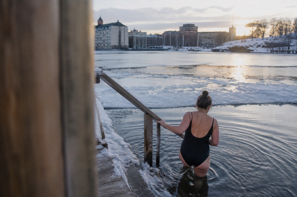 Person stepping from a snowy sauna deck into dark fjord water between ice rims