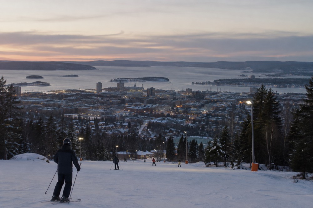 Skiers on a floodlit slope above the lights of a city at dusk