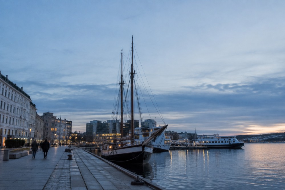 A wooden sailing ship and small ferries moored along a city waterfront promenade