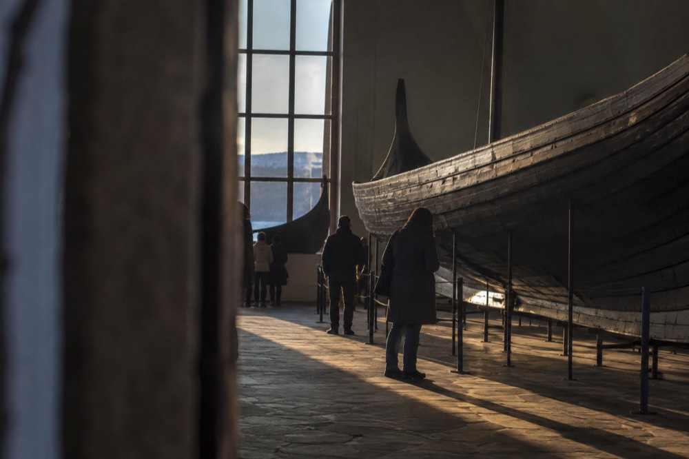 Visitors in a dim museum hall on Bygdøy studying old wooden expedition boats