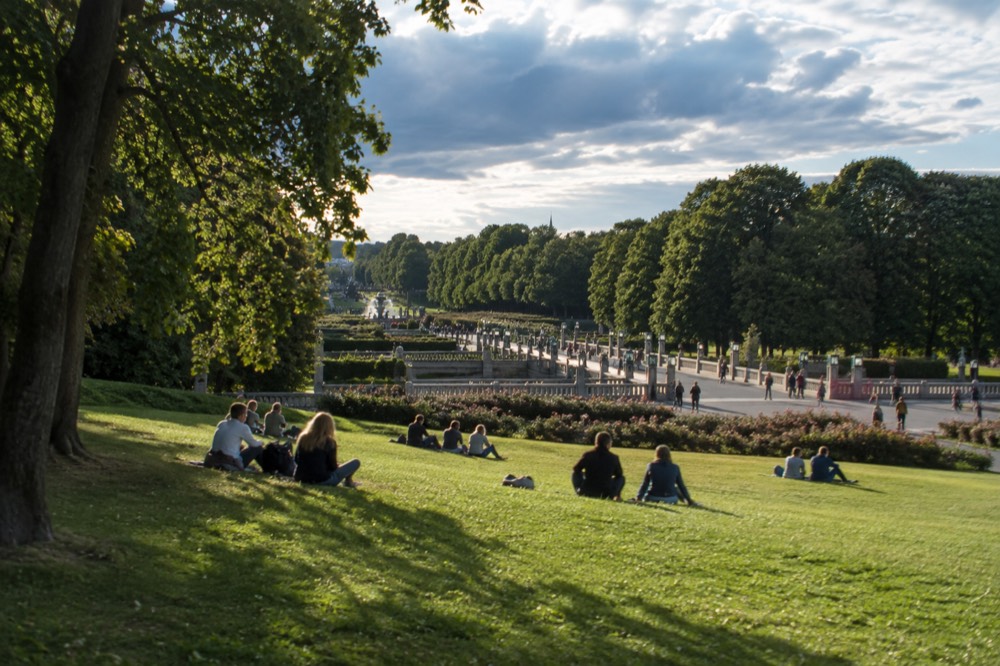 Visitors lounging on the lawns beneath tall trees at the edge of Frogner Park